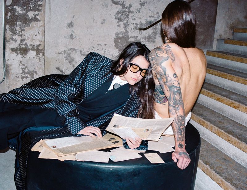 Two people in a dimly lit interior with a concrete wall and stairs in the background, papers scattered on a black cylindrical ottoman.