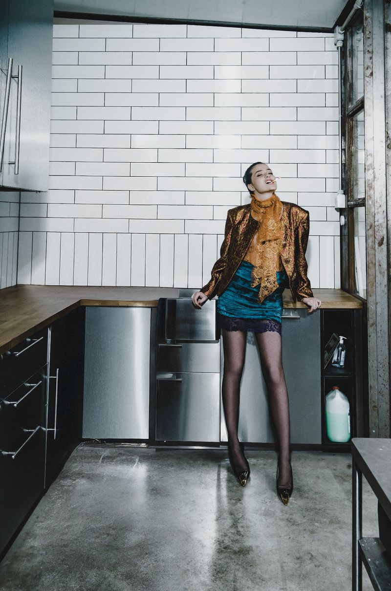 A stylish adult woman in gold jacket and blue skirt poses in a kitchen with white brick backsplash, stainless steel appliances, and concrete floor.