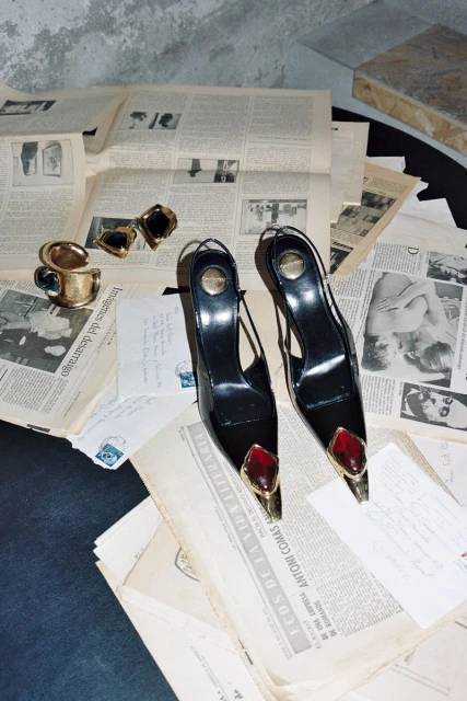 Fashion still life features pair of black slingback heels with gold and ruby accents, placed amid scattered vintage newspapers and gold jewelry.