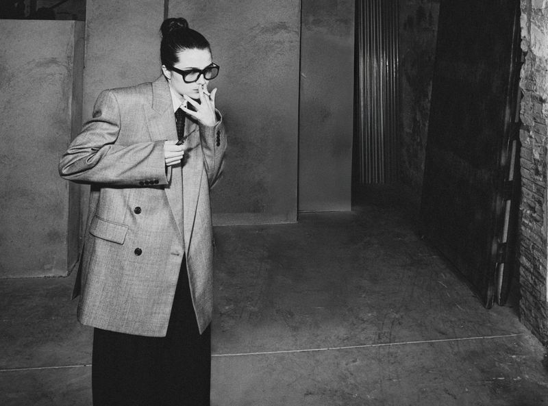 Monochrome portrait of a stylish adult female in an oversized blazer, smoking indoors with harsh lighting, posing in front of a door.