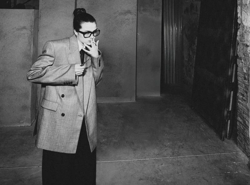 Monochrome portrait of a stylish adult female in an oversized blazer, smoking indoors with harsh lighting, posing in front of a door.