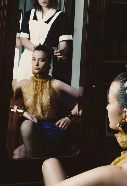 Woman in gold halter top is reflected in mirror as another woman adjusts her hair indoors with dark wood and white surfaces.