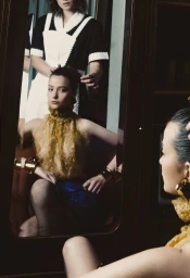 Model Blanca Soler seated before a mirror wearing a sheer gold textured top and bold gold cuffs, with another person styling her hair.