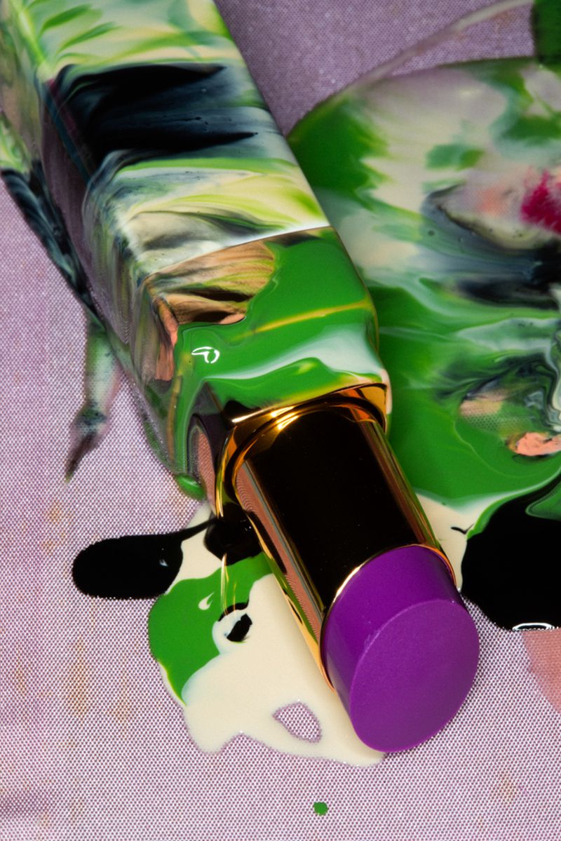 A close-up studio shot displays an open lipstick tube with a purple tip, surrounded by colorful green, white, and black cosmetic drips.