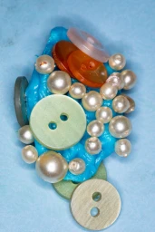 Close-up of a blue glossy gel blob embedded with various pearl beads and multi-colored plastic buttons on a blue background.