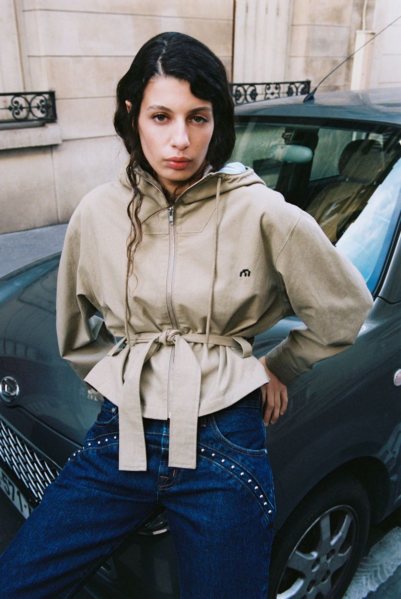 A woman in a beige jacket and dark denim poses leaning against a teal car in an urban setting with a neutral-toned building backdrop.