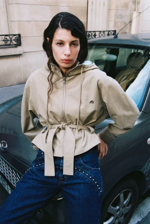 A woman in a beige jacket and dark denim poses leaning against a teal car in an urban setting with a neutral-toned building backdrop.