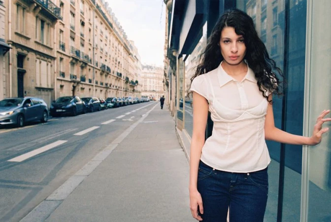 Woman in white shirt and jeans poses on city sidewalk next to building with cars parked along the street in the background.