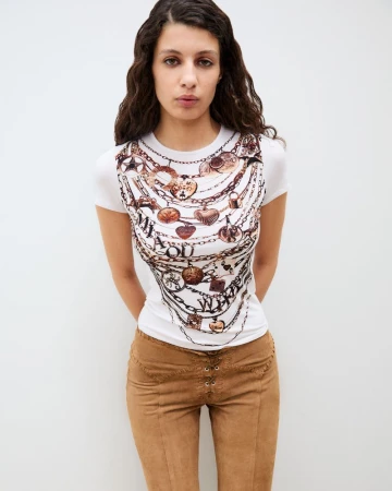A slender adult woman poses indoors wearing a white tee with a printed design and tan suede pants against a white wall.
