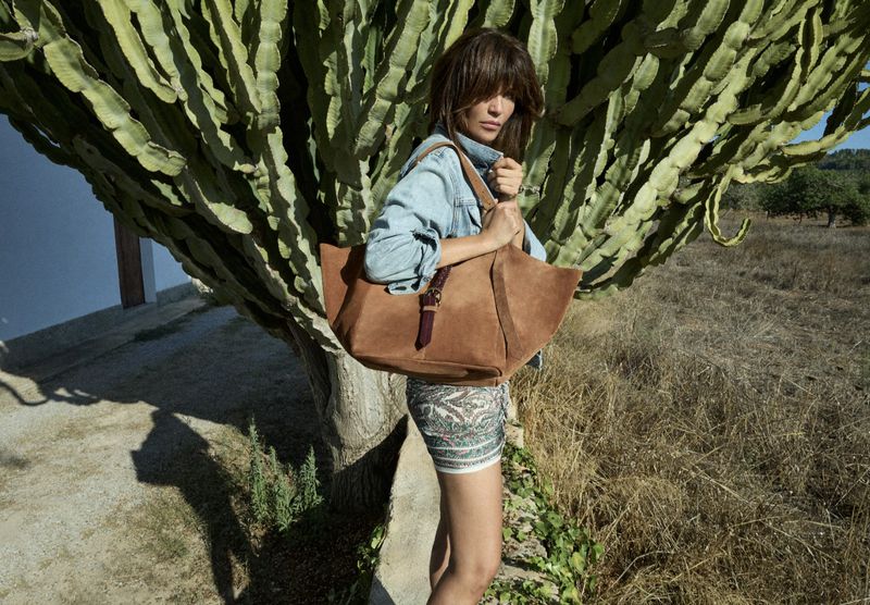 A fashionable woman with a brown suede tote bag standing outdoors in front of a large cactus, wearing a denim jacket and patterned skirt.