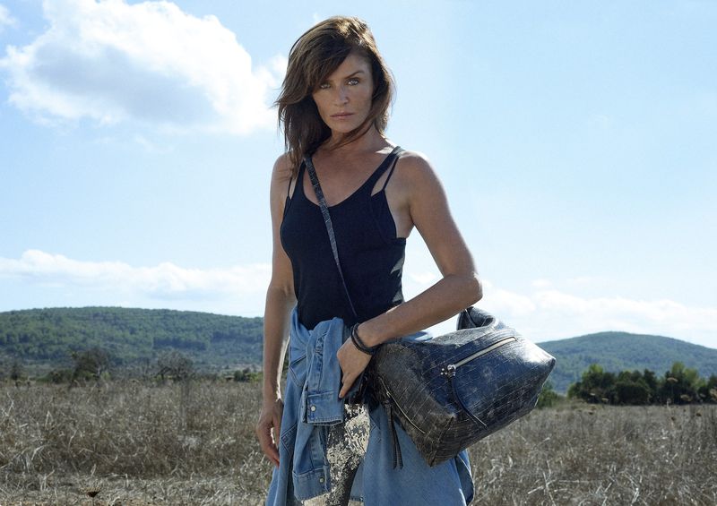 Woman standing outdoors wearing black tank top, denim jacket, carrying a large bag against a backdrop of sky and grassy hills.