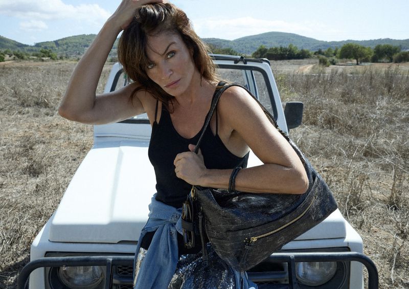 Adult woman poses with an over-the-shoulder black hobo bag, wearing a black top and jeans with a vintage white SUV in a field.