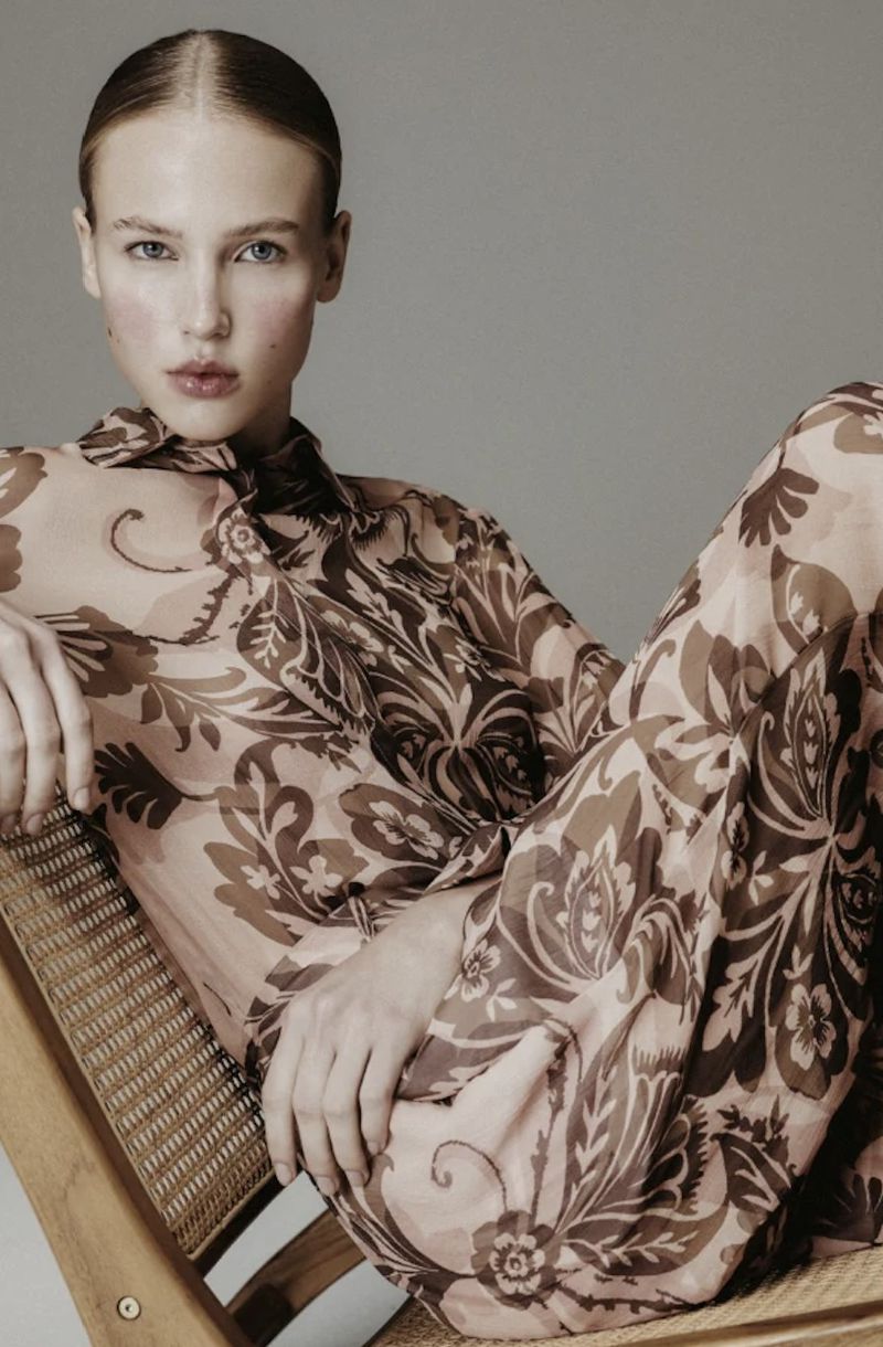 Blonde woman in floral patterned top and pants poses in a studio on a wooden chair, gazing at the camera with neutral expression.