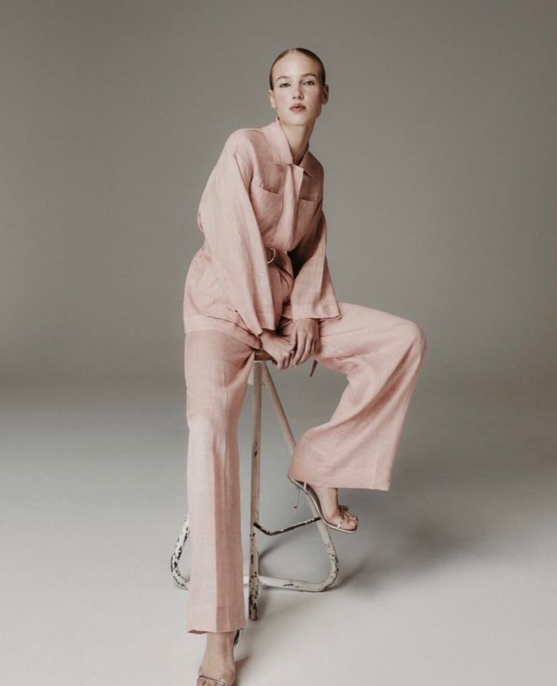A fair-skinned woman sits on a stool wearing a blush-colored linen suit with wide-legged pants, in a clean photo studio setting.