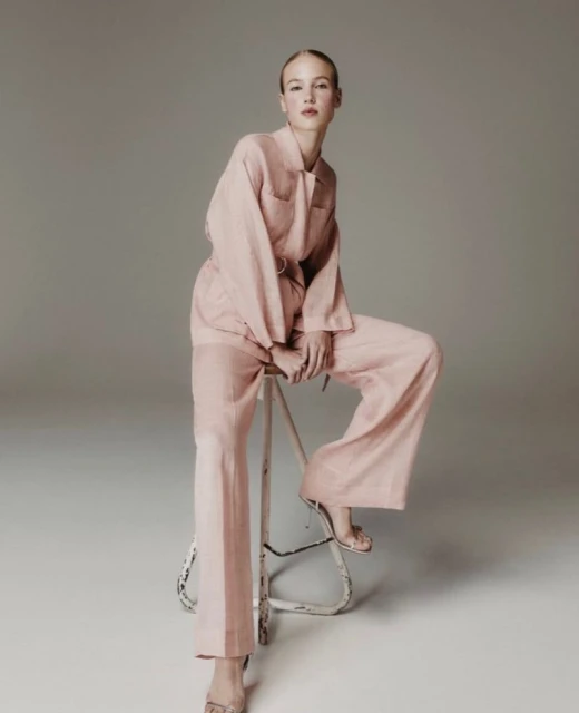A fair-skinned woman sits on a stool wearing a blush-colored linen suit with wide-legged pants, in a clean photo studio setting.