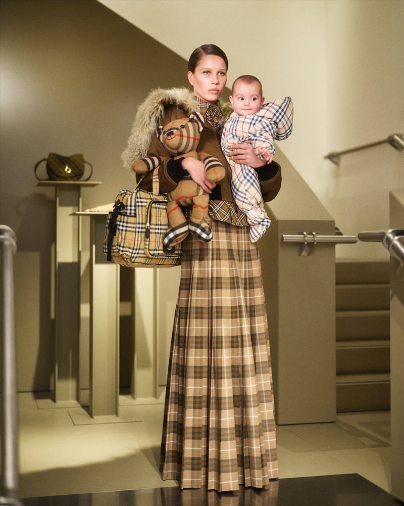 Woman holding baby wearing Burberry plaid outfit with plaid diaper bag and teddy bear; beige, taupe, and brown tones dominate.
