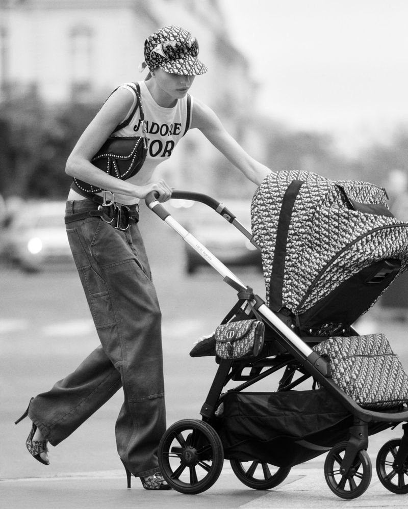 A woman in a Dior hat and jeans pushing a Dior stroller on a city street, in a black and white fashion shot. She wears heels and a bag.