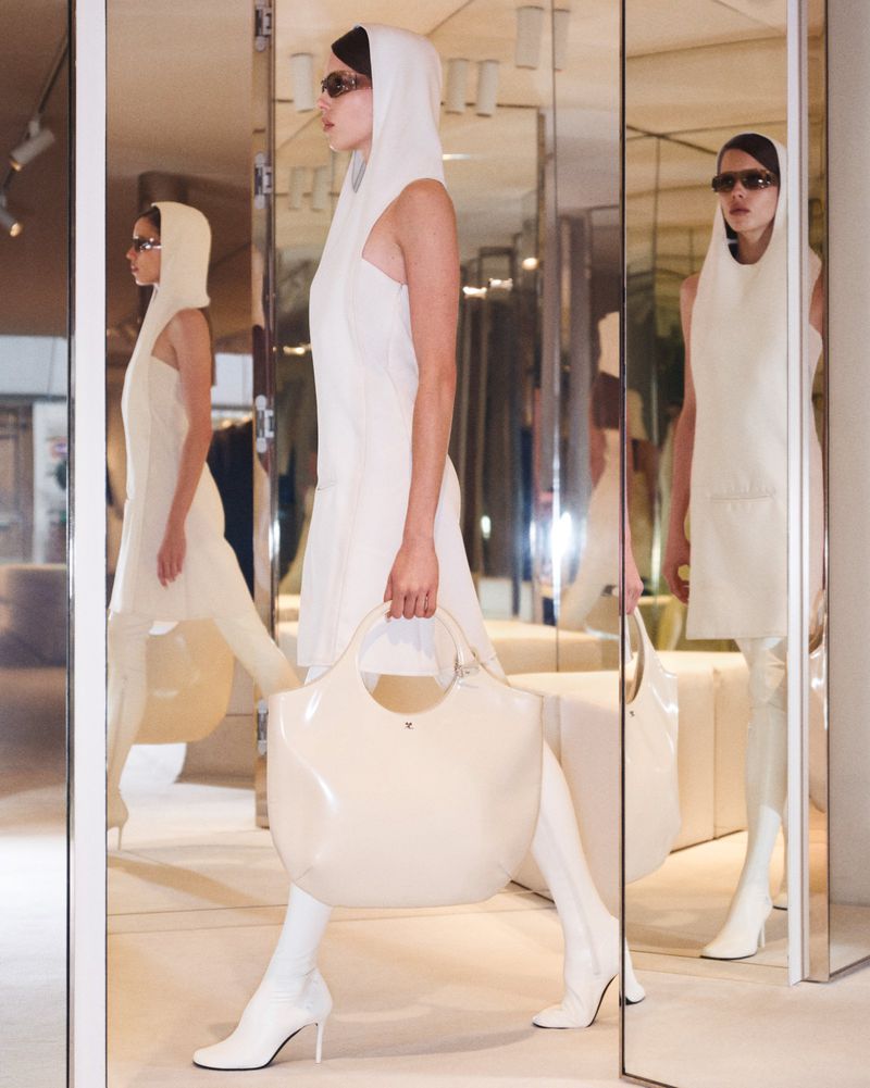 A woman in a white hooded dress and matching boots, carrying a white handbag, stands in front of a mirror, showcasing a fashion-forward look.