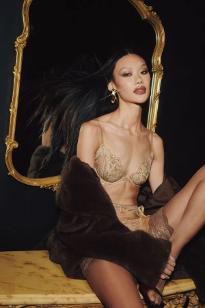 Model wearing gold lace lingerie and a brown fur coat seated before an ornate gold-framed mirror.