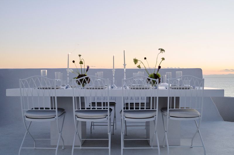Outdoor dining table setup with white chairs, candles, floral arrangements, and glassware, set against an ocean and sky backdrop at sunset.