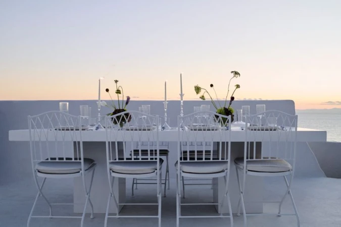 Outdoor dining table setup with white chairs, candles, floral arrangements, and glassware, set against an ocean and sky backdrop at sunset.