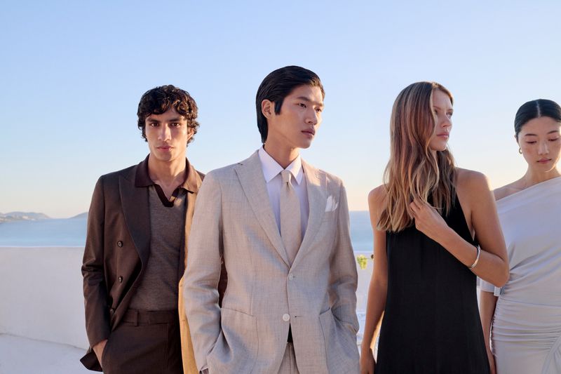Four fashion models pose outdoors on a sunny day showcasing a light linen suit, a brown suit, and two dresses in black and white.