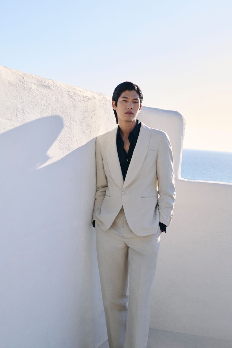 An adult male with dark hair wearing a tan suit and black shirt stands against a white wall with a view of the sea and clear sky.