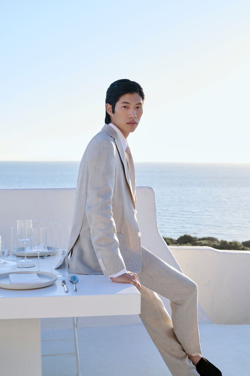 An Asian man wearing a light-colored suit is sitting on a white table outdoors with the ocean in the background. The scene is bright and airy.