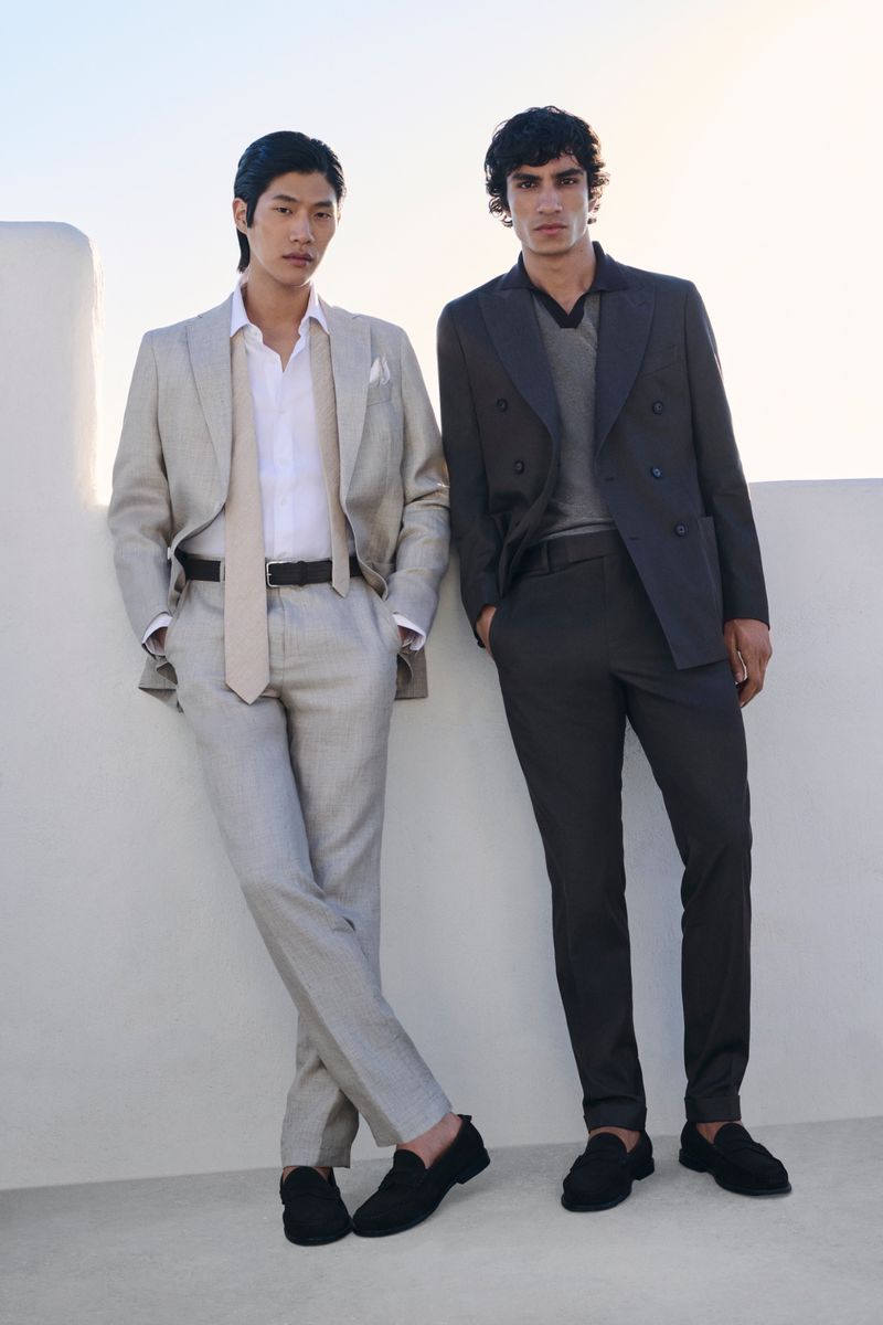 Two adult men pose in suits against a white stone wall in natural light, showing fashion-forward menswear in a clean, outdoor setting.