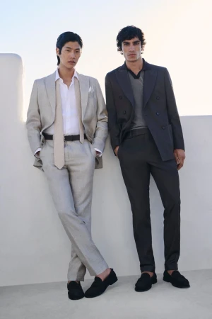 Two adult men pose in suits against a white stone wall in natural light, showing fashion-forward menswear in a clean, outdoor setting.