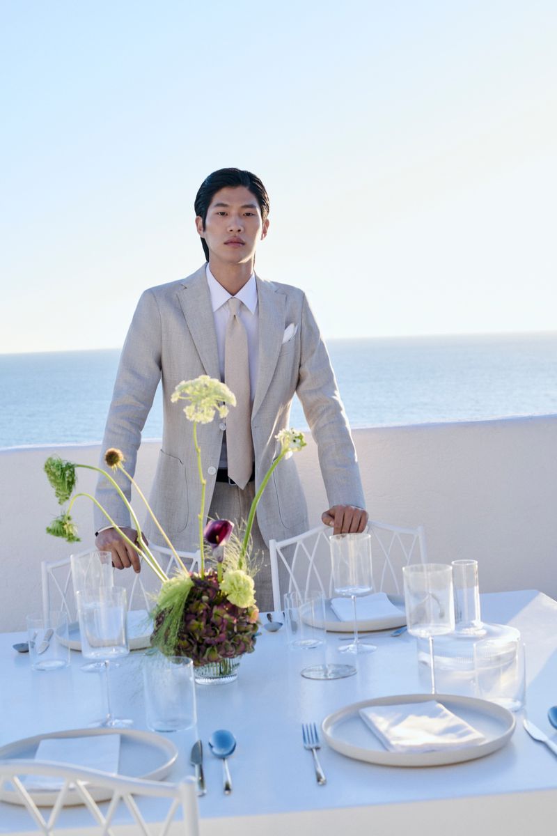 Asian man in a light suit standing behind a white table set with dinnerware, with the ocean and blue sky in the background.