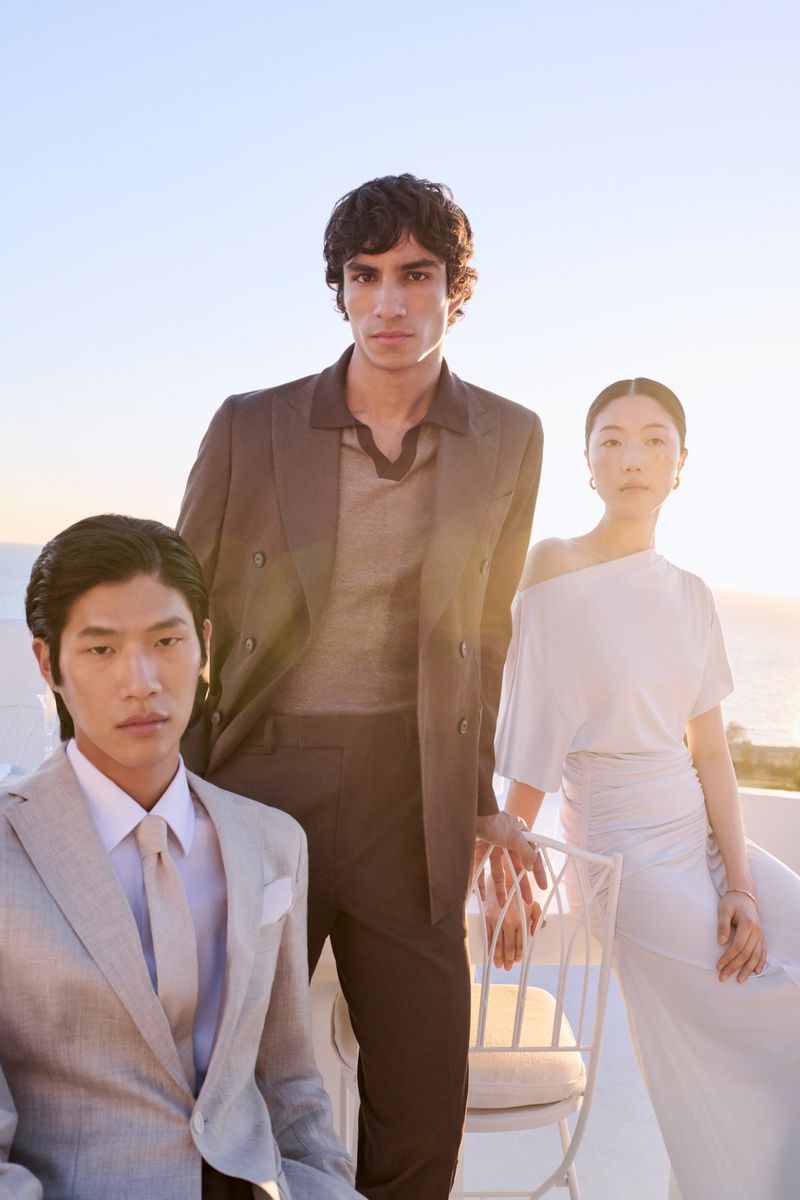 Fashionable group portrait features three young adults in suits against a bright blue sky backdrop with subtle coastal horizon.