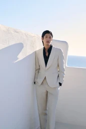 Model in cream suit and black shirt leaning against white stucco wall with sea view