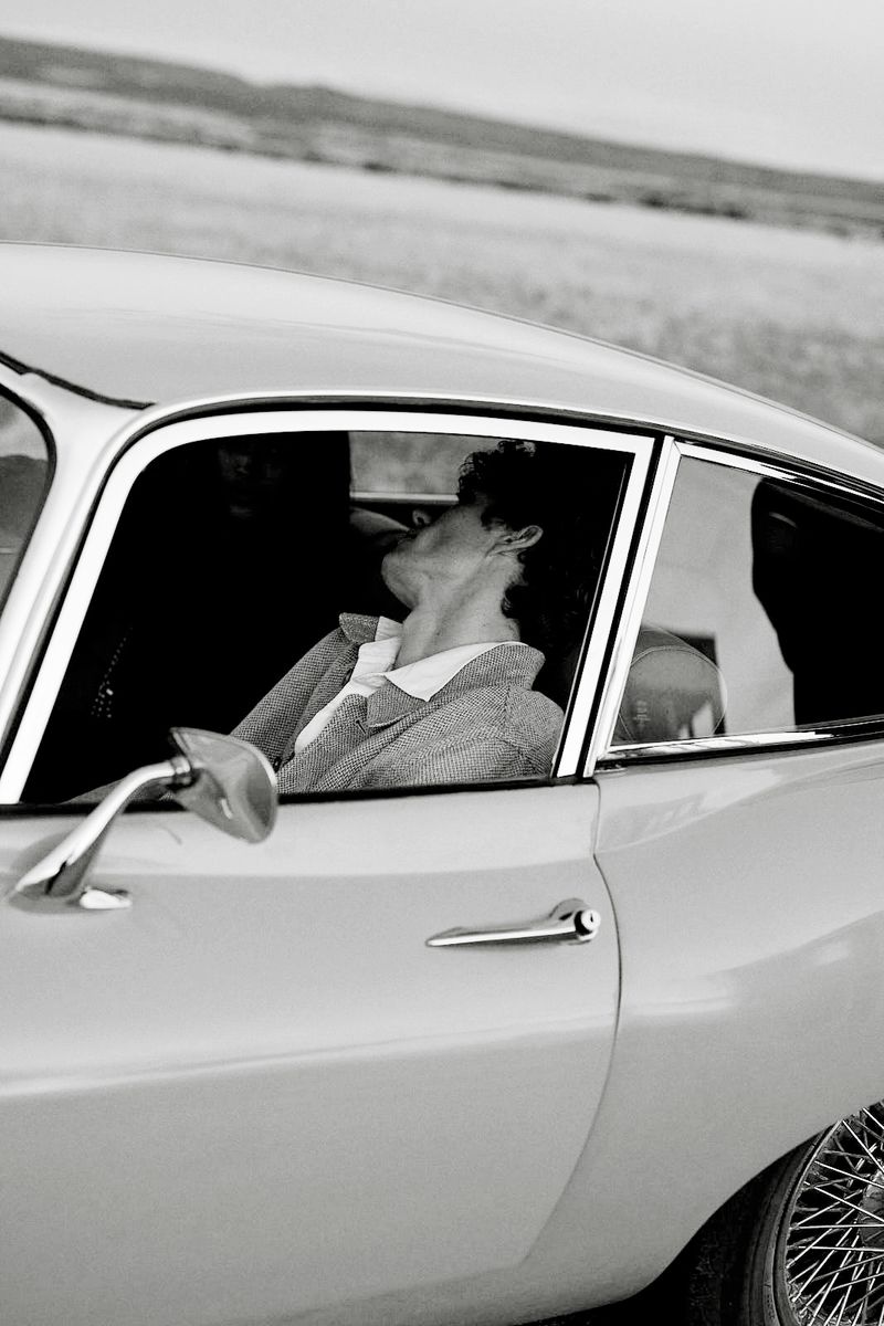 Monochrome portrait of an adult woman in a vintage car, with a glimpse of an outdoor landscape in the background, evoking a timeless feel.