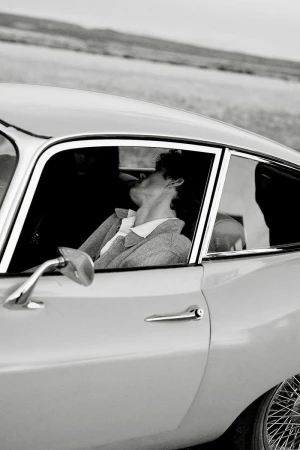Monochrome portrait of an adult woman in a vintage car, with a glimpse of an outdoor landscape in the background, evoking a timeless feel.