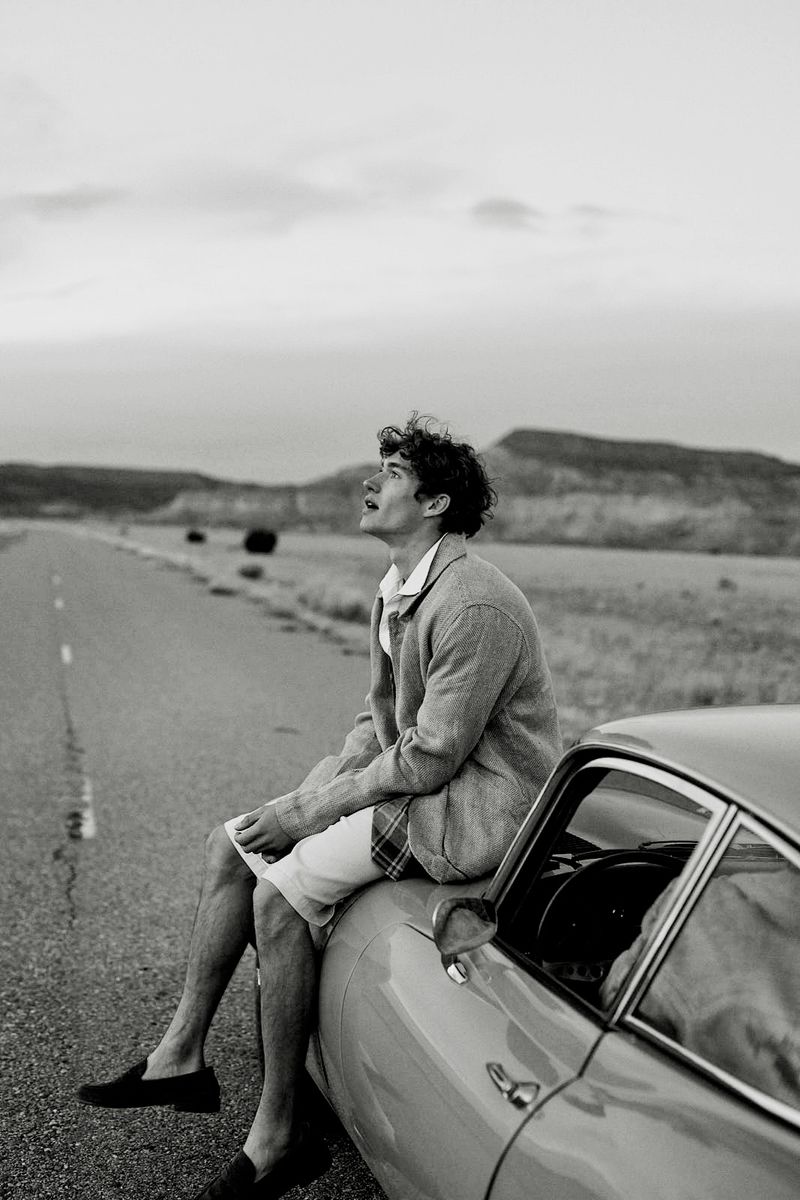 Black and white photograph of a curly-haired man sitting on a vintage car on an open road, gazing upwards with mountains in the background.