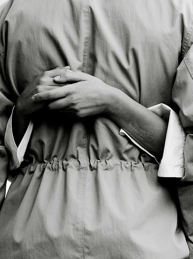 Close-up shot of a person wearing a jacket, their hands clasped behind their back, in a monochrome photo studio setting, with soft lighting.