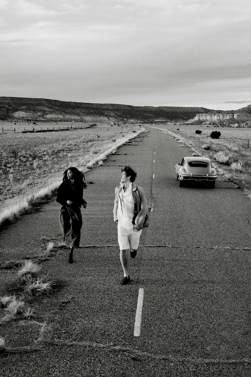 Two people run towards the camera on a long highway in a monochrome photo, with a vintage car visible in the distance.