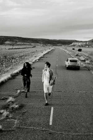 Two people run towards the camera on a long highway in a monochrome photo, with a vintage car visible in the distance.