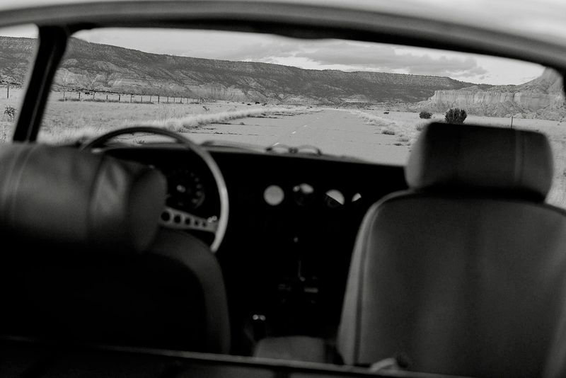 Black and white interior view of a vintage car aimed towards a desert landscape, featuring a straight road and distant mountains.