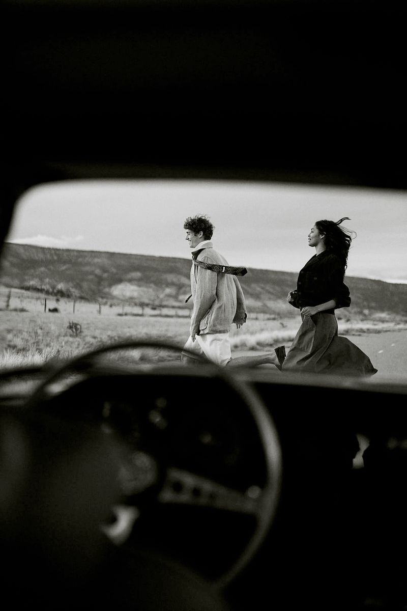A man and woman running across a rural road as viewed through the windshield of a vintage car in black and white.
