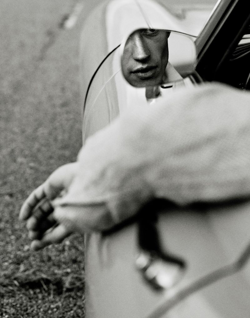 A man's face is reflected in the side mirror of a vintage automobile in this grayscale photograph with hand and car door.