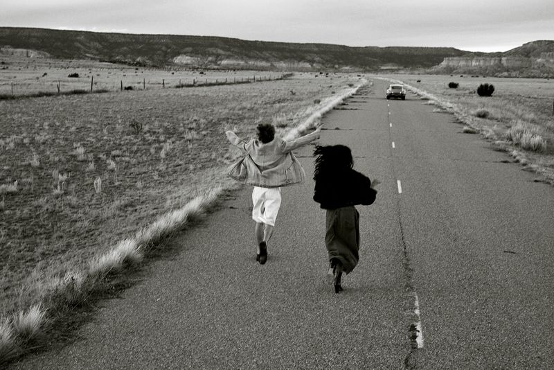 Two figures, a male and female, run away from the camera down a deserted road with a vintage car in the distance, in a monochrome landscape.