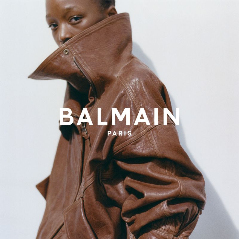 Fashion-forward portrait of a Black woman showcasing a voluminous brown leather jacket with the Balmain logo against a plain background.