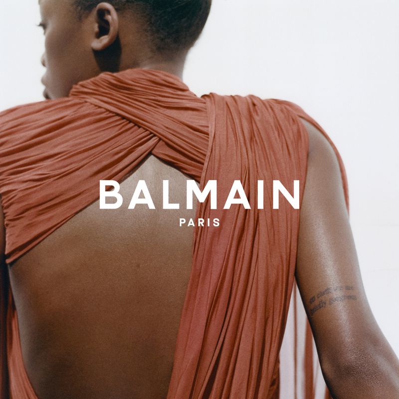 Model wearing a draped terracotta backless dress against a plain white background with bold Balmain Paris white typography centered across the frame.