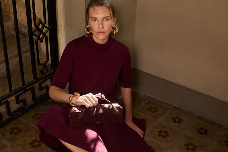 Woman in burgundy dress sitting with burgundy handbag on patterned tile floor. Gold jewelry, indoor setting. Fashion and style.