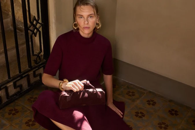 Model in burgundy knit outfit sitting on patterned tile floor holding a structured handbag