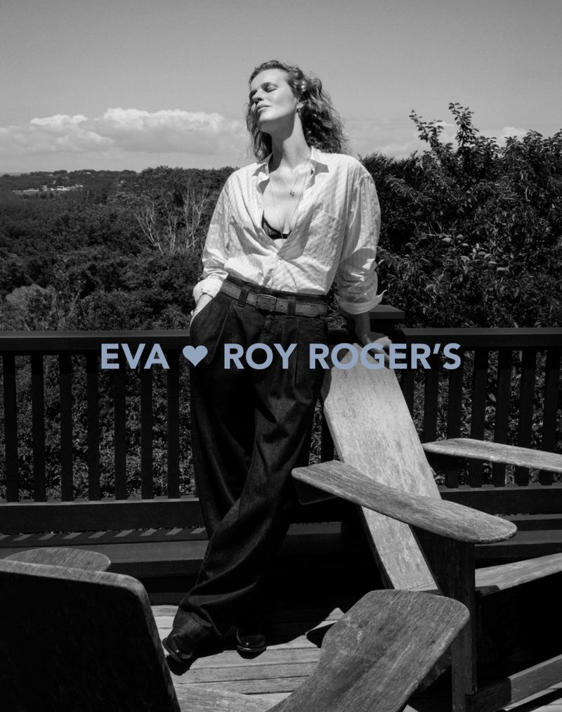 A woman poses on a balcony with a rural vista in black and white. She wears an unbuttoned shirt and wide-leg pants, with a relaxed expression.