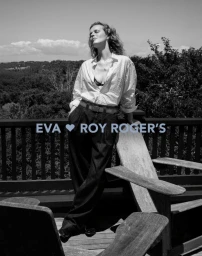 Model Eva Herzigova leaning against a wooden deck chair outdoors, wearing a white button-down shirt and dark trousers for Roy Rogers S/S 2026.