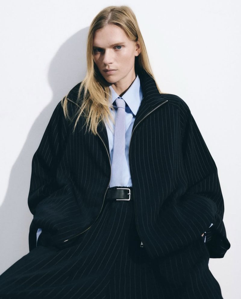 A young adult with long blond hair poses indoors, wearing a pinstripe jacket, light blue shirt and tie, in a fashion editorial shot.